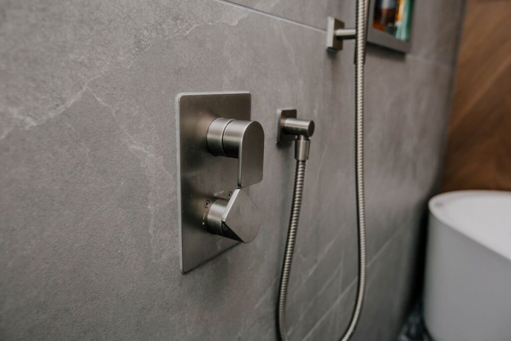 Close up of a brushed gunmetal thermostatic shower valve and handheld holder on grey stone look porcelain wall tile