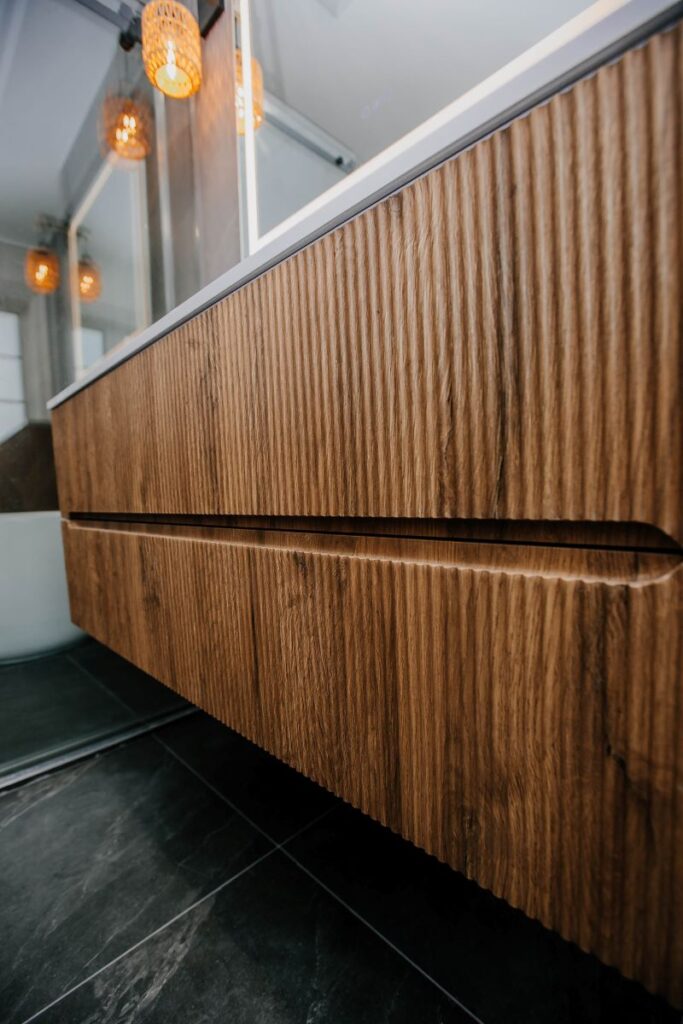 Detail of a fluted oak floating vanity with warm amber pendant lights reflected in the LED mirror above