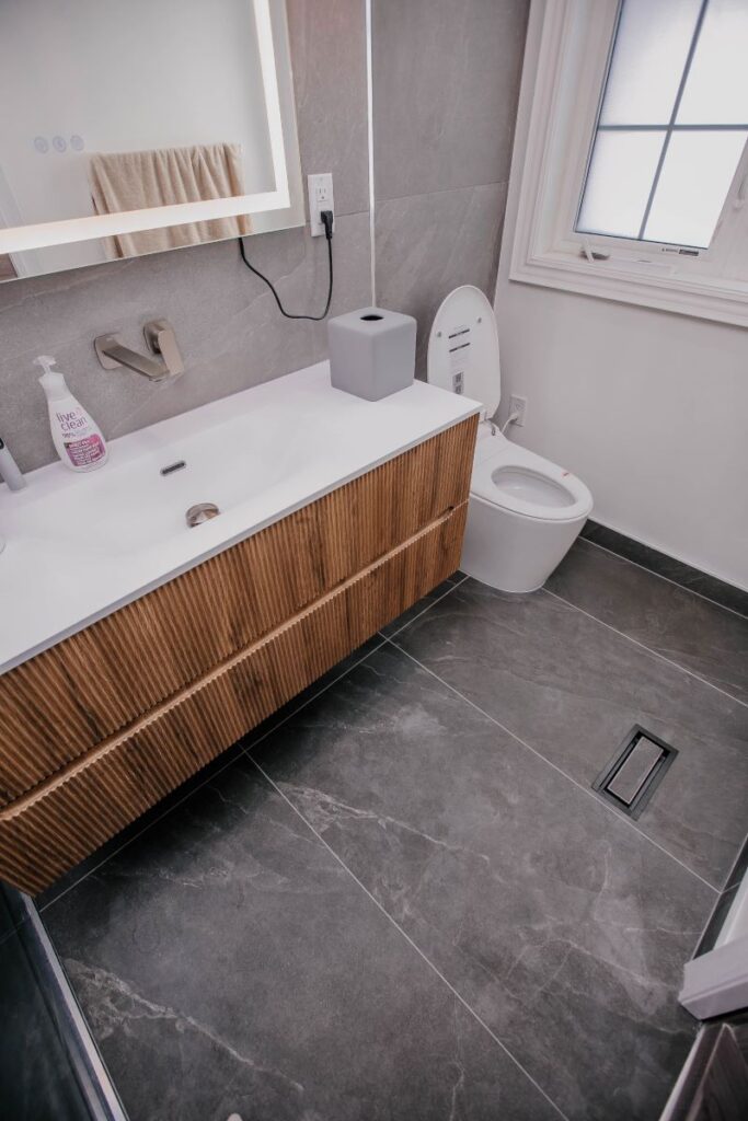 Fluted oak floating vanity with integrated white basin and brushed nickel wall faucet beside a smart toilet on charcoal slate look porcelain flooring