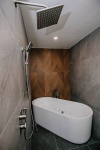 Square brushed nickel rain shower and handheld over a freestanding soaking tub against a walnut chevron feature wall