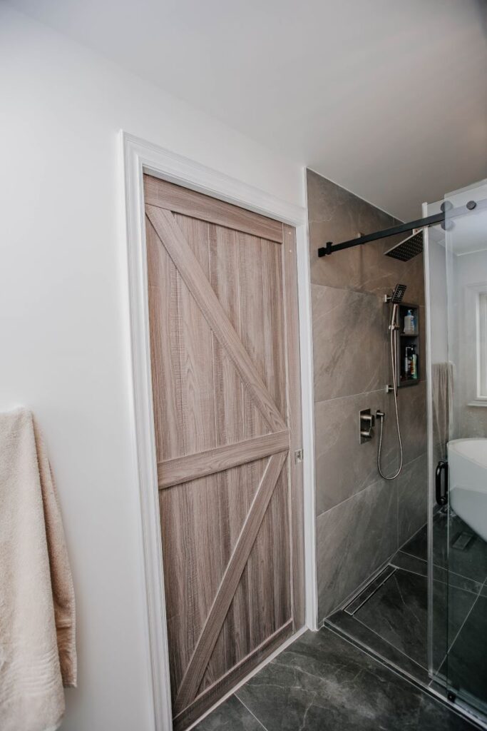 Wood look sliding barn door beside a grey porcelain walk in shower with brushed nickel rain head and frameless glass
