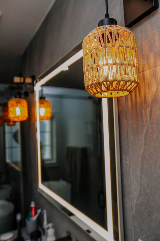 Woven rattan pendant light glowing warm amber above a frameless LED mirror on a grey stone look porcelain wall