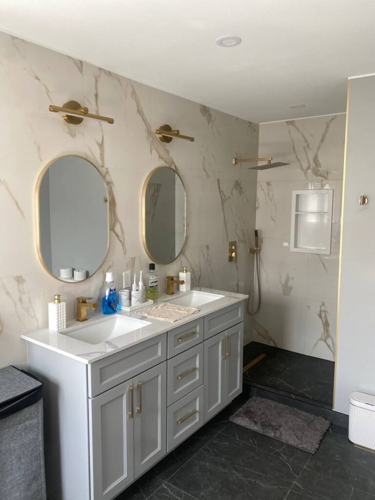 Close up view of the double vanity with grey cabinetry white countertop gold hardware and two oval mirrors with gold frames Wall mounted gold light fixtures above the mirrors