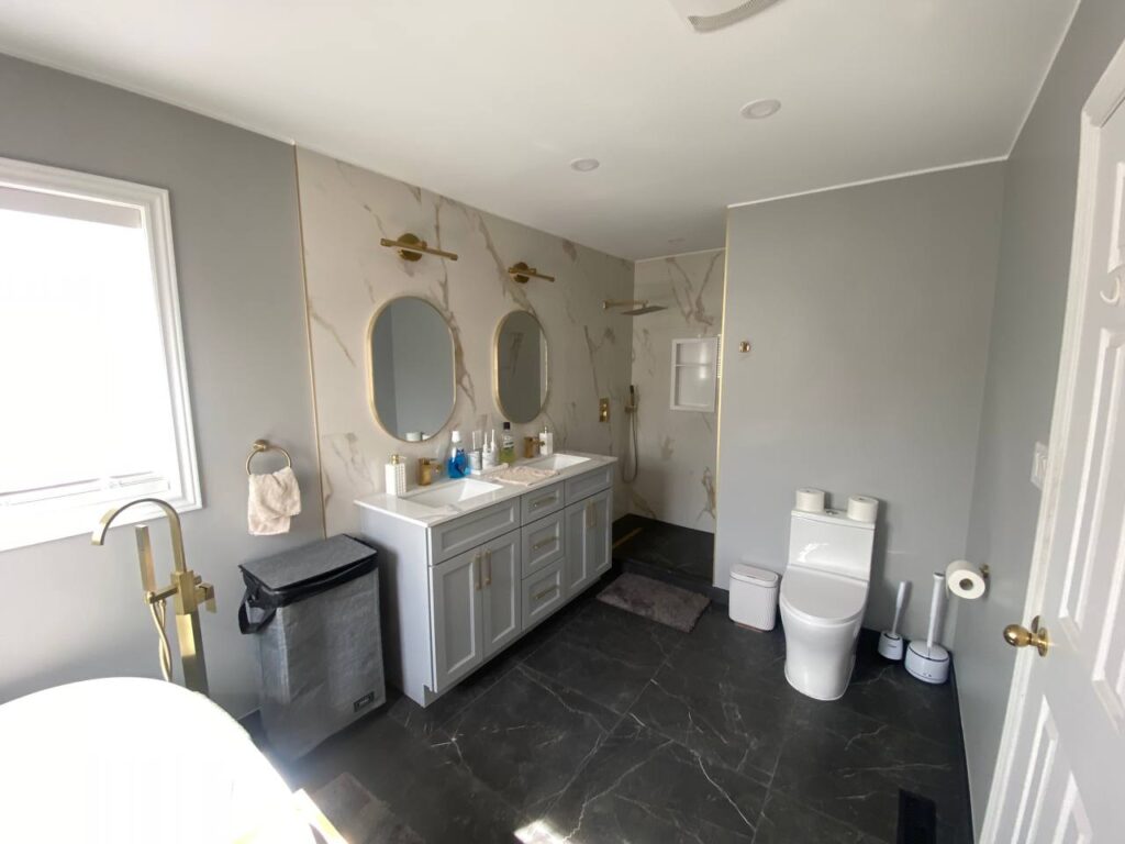 Panoramic view of the renovated bathroom showcasing the shower area double vanity toilet and part of the bathtub The design highlights the grey and gold color scheme with marble effect tiles