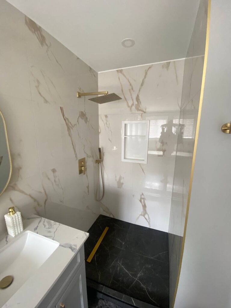 Walk in shower with marble effect tiles and gold fixtures next to double vanity with grey cabinetry white countertop and gold hardware Two oval mirrors with gold frames and wall mounted light fix