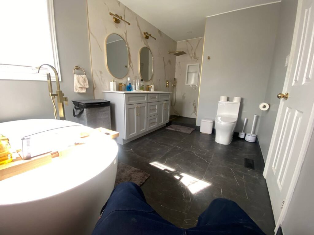 Wide angle view of the renovated bathroom featuring walk in shower double vanity bathtub and toilet. The design includes grey cabinetry gold fixtures and dark marble floor tiles
