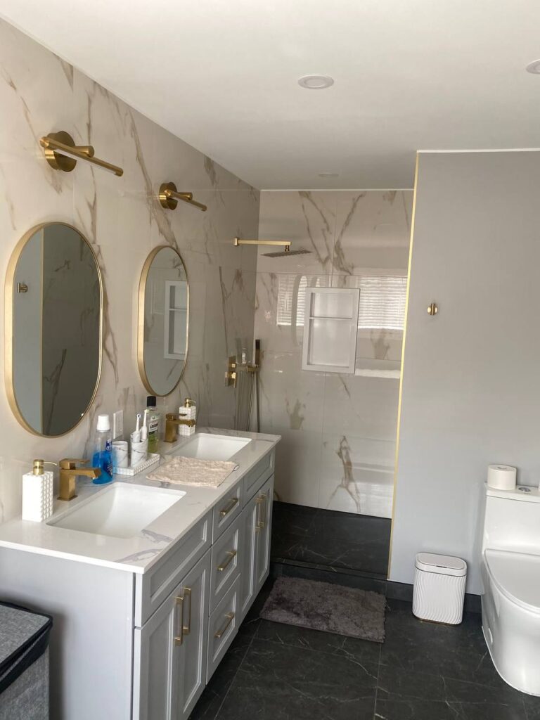 modern bathroom with double vanity gold fixtures and marble tiles There are two oval mirrors above the sinks gold sconces on the wall and large walk in shower with rain shower head