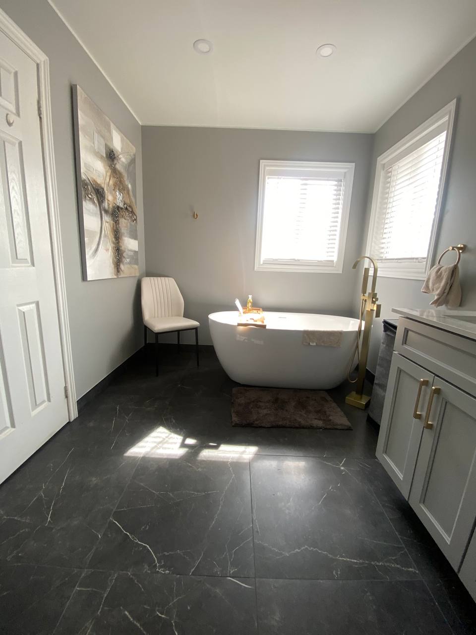 bathroom remodel in toronto: modern bathroom with freestanding white bathtub gray wall and dark tile floor large abstract painting on the wall bathtub nish with white blinds gold fixtures add touch of luxury Aurora