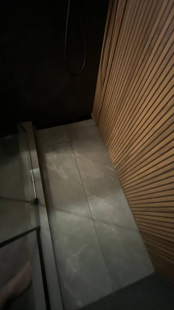 A top down view of the shower floor, featuring grey stone look tiles. The bottom of the vertical wood slat accent wall is visible on the right. Part of the glass enclosure is also in the frame