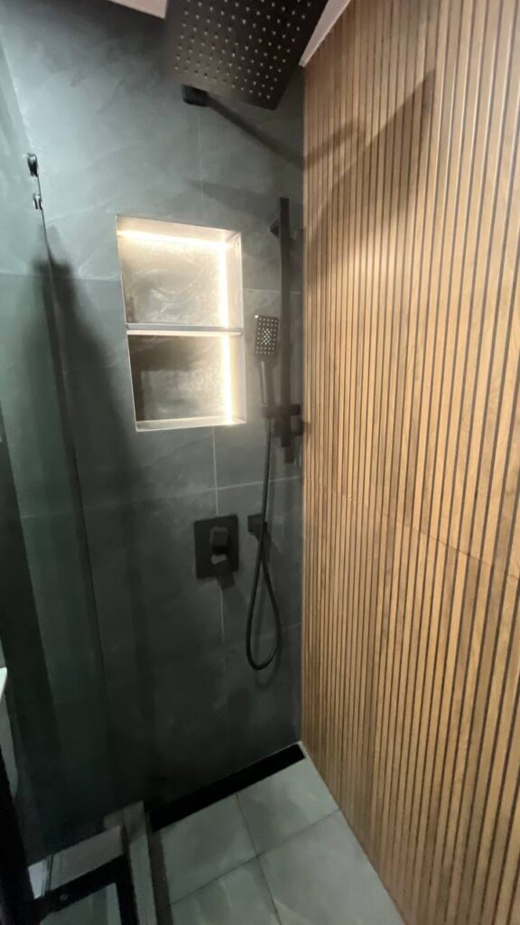 Close up of a walk in shower with a wood slat accent wall and a recessed niche with integrated LED lighting. Matte black shower fixtures are visible
