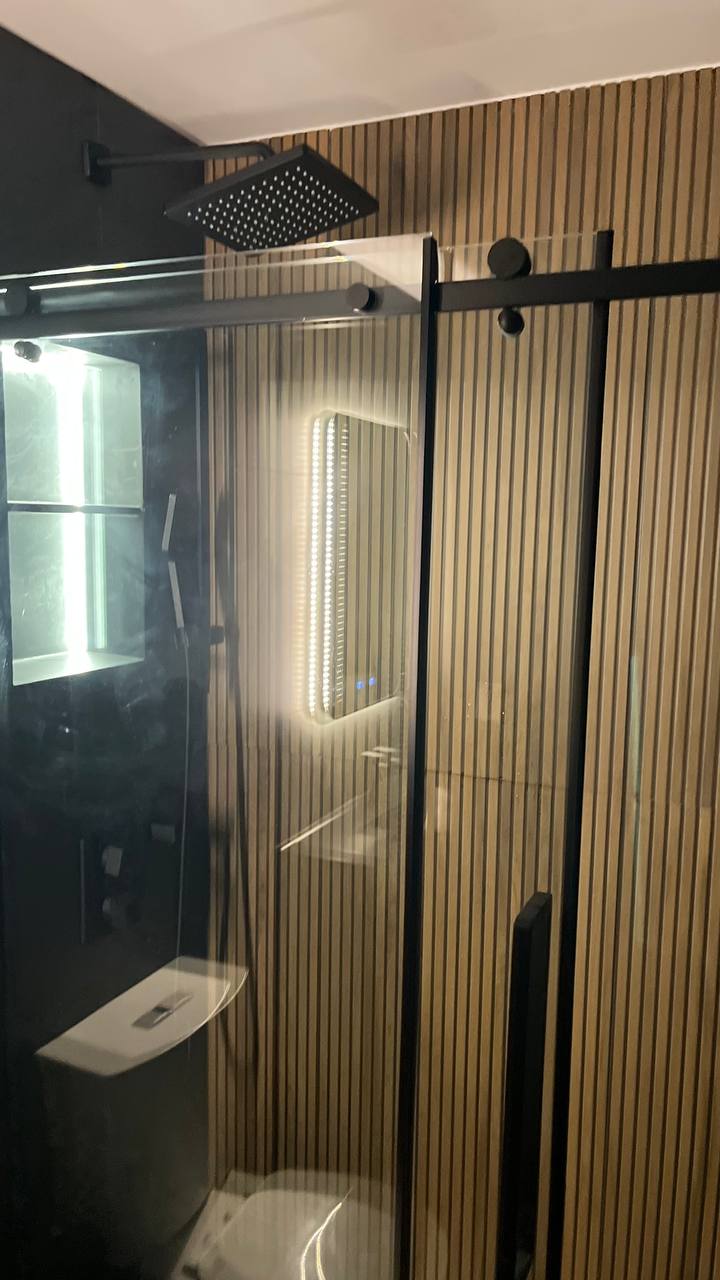 Close up of the shower system. A black square rain showerhead, a glass door with a black handle, and the wood slat wall are visible. Inside the shower, a built in vertical niche is illuminated