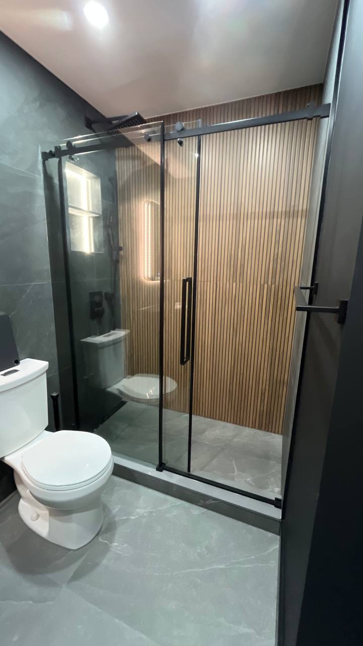 Bathtub to shower. Frameless sliding glass shower door with matte black hardware in a modern bathroom, with a wood slat accent wall inside