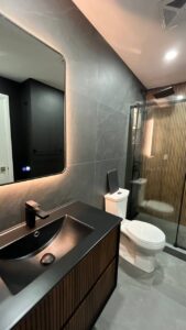 Wide view of a modern dark bathroom showing the floating wood vanity, LED mirror, white toilet, and walk in shower with a wood slat wall