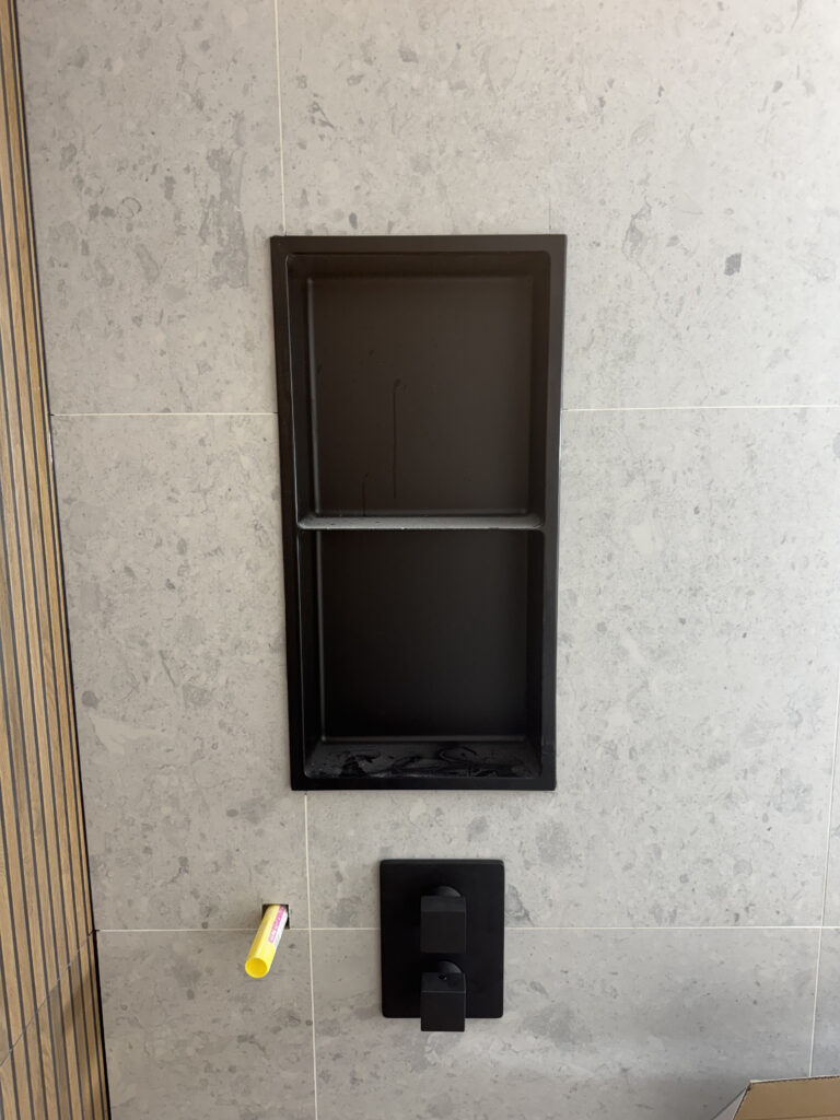 Bathroom renovation construction phase. Close up a matte black built in shower niche and matte black shower valve controls on the light gray tile wall. Details of the modern bathroom design