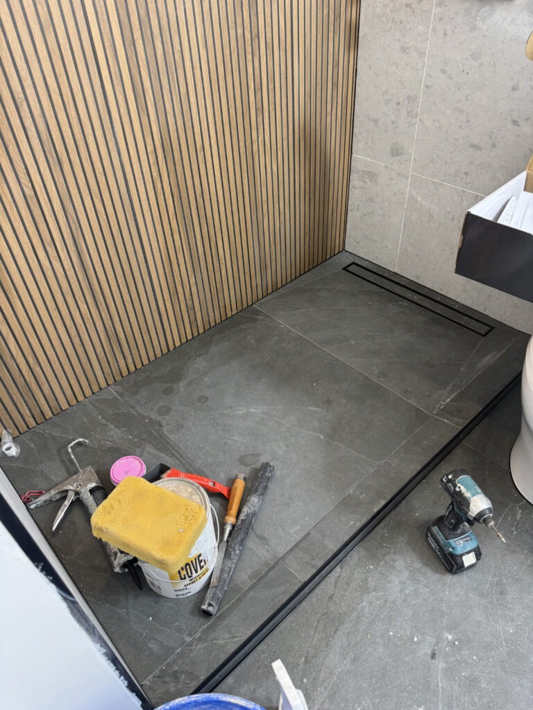 Bathroom renovation construction phase. Close up of the walk in shower area. Dark gray floor tile with black linear drain. Tools and materials visible. Shows the wooden slat accent wall