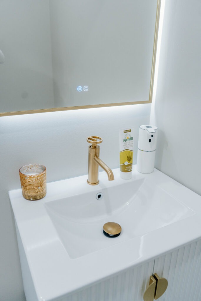 Close up detail of the brushed gold faucet and integrated sink on the floating vanity, with the touch controls of the LED mirror visible