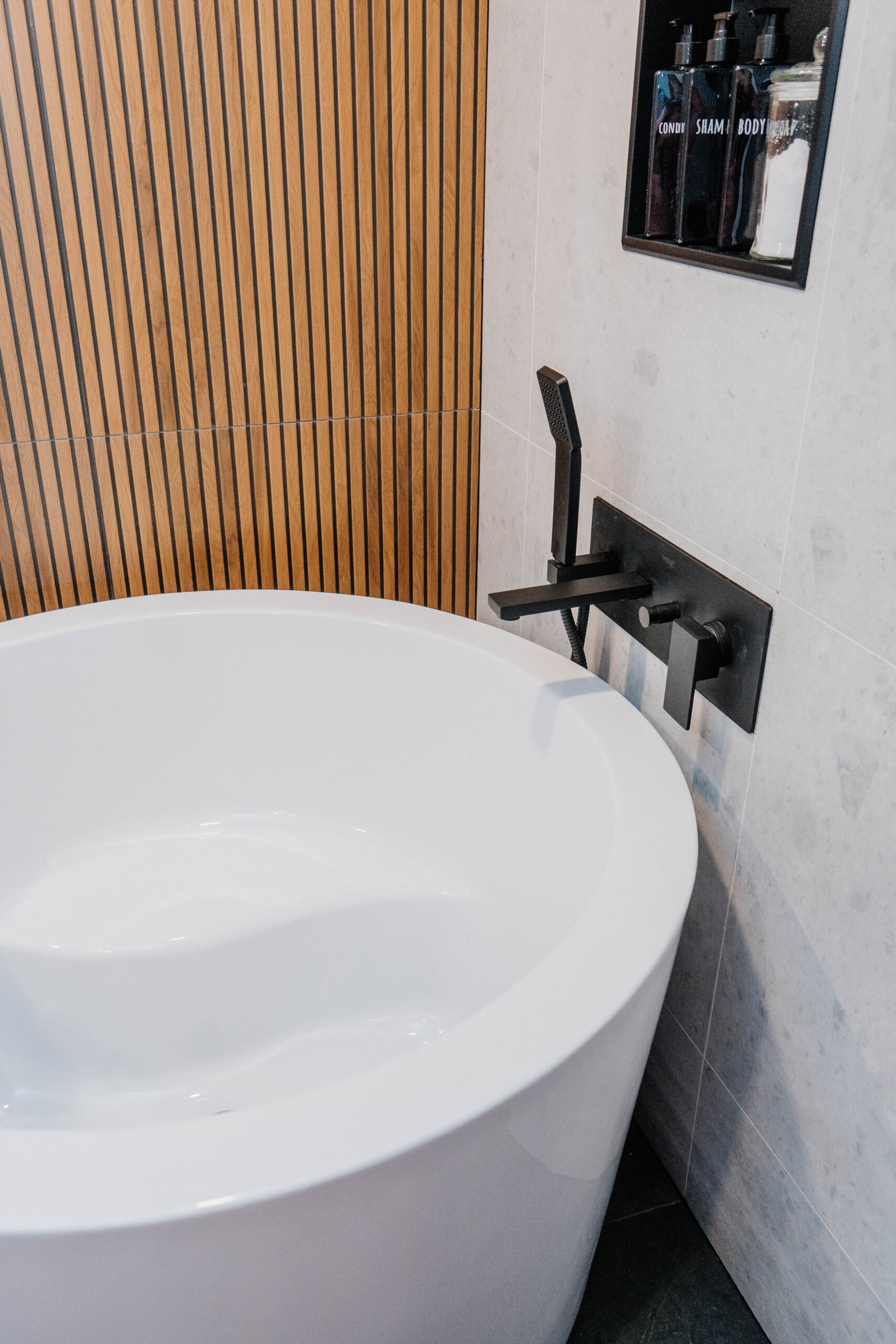 Close up sleek white freestanding circular bathtub with a matte black wall mounted faucet. Gray tile and built in niche visible. Luxury bathroom renovation, GTA