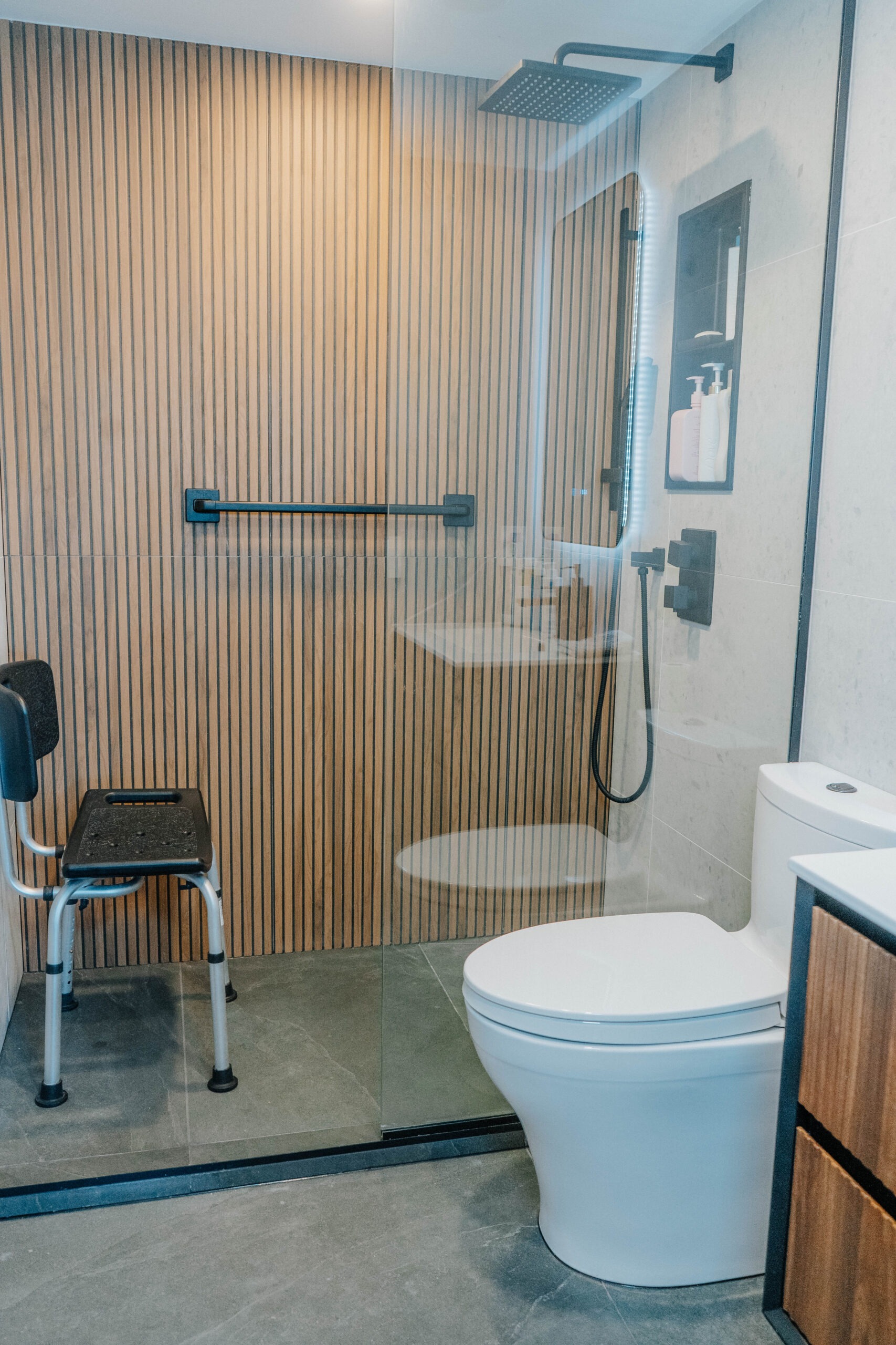 Curbless walk in shower with matte black fixtures and a wooden accent wall. White toilet and shower chair are visible. Accessible bathroom renovation, modern design GTA