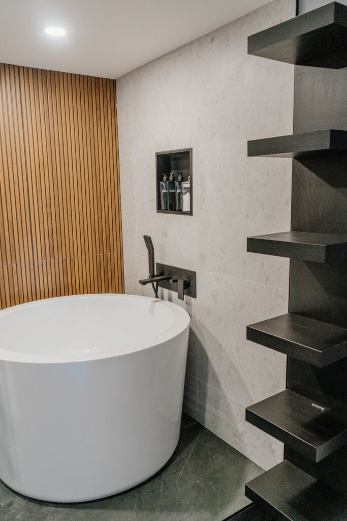 Freestanding bathtub, black floating shelves, and a wooden slat accent wall. Modern spa style bathroom renovation