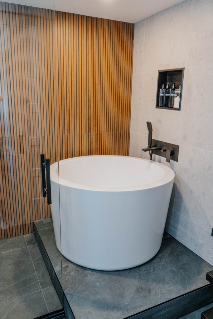Freestanding bathtub on a dark podium next to the wooden slat wall. Matte black wall mounted faucet and built in storage shelf. Modern bathroom design, Magic Reno
