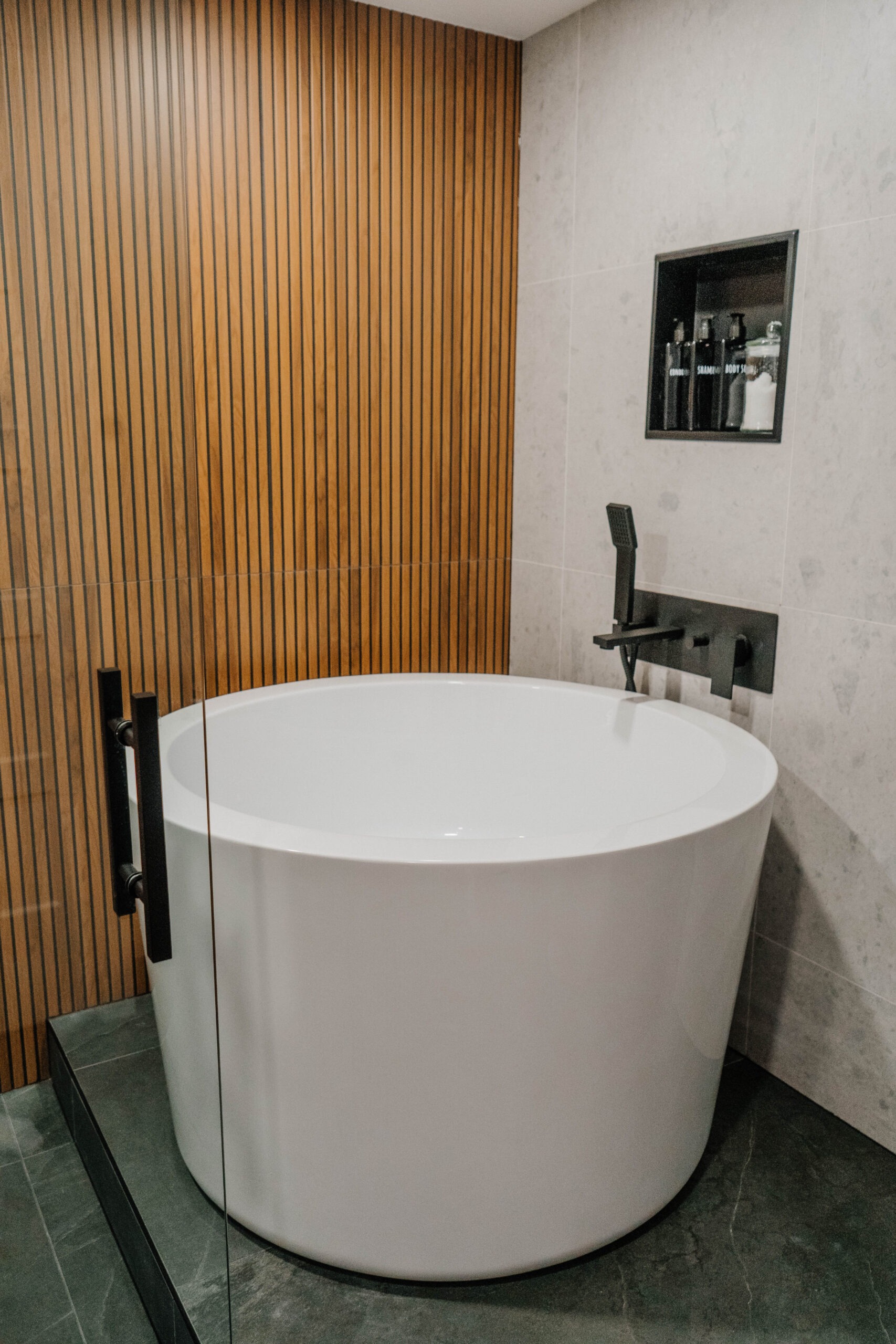 Freestanding circular bathtub on a raised dark podium, enclosed by a glass partition. Wooden slat accent wall provides warmth. Modern bathroom renovation by Magic Reno
