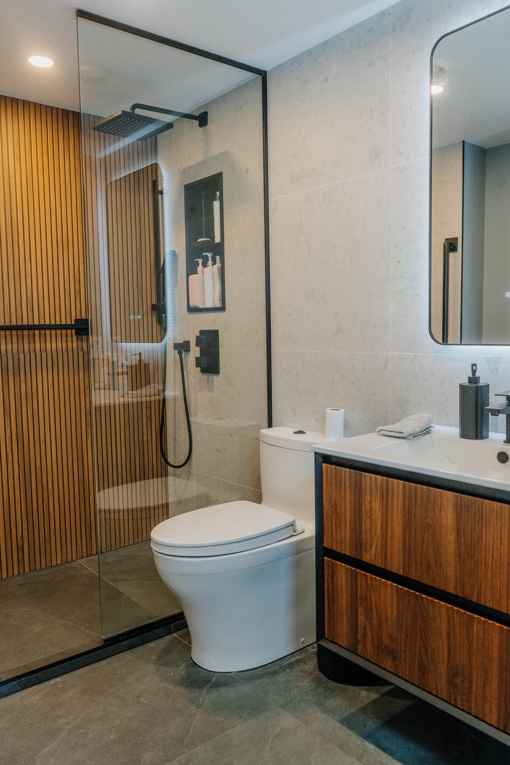 General view of a modern bathroom renovation. Design featuring wood slats in the shower, glass partition, white toilet, and wooden vanity. Modern bathroom design by Magic Reno in TorontoGTA