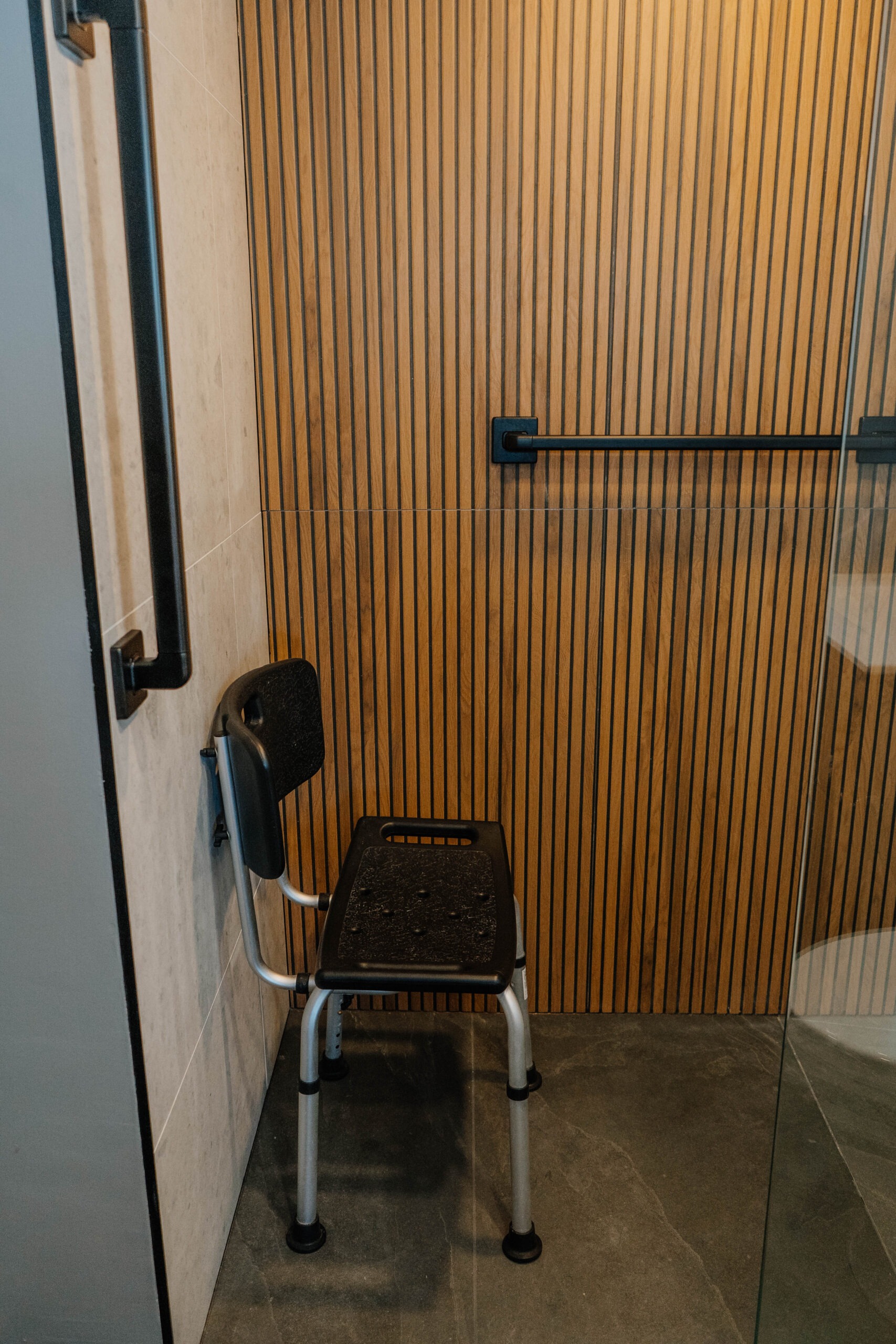 Shower detail wooden slat accent wall, black towel bar, and a transfer benchshower chair. Accessible bathroom renovation, modern design