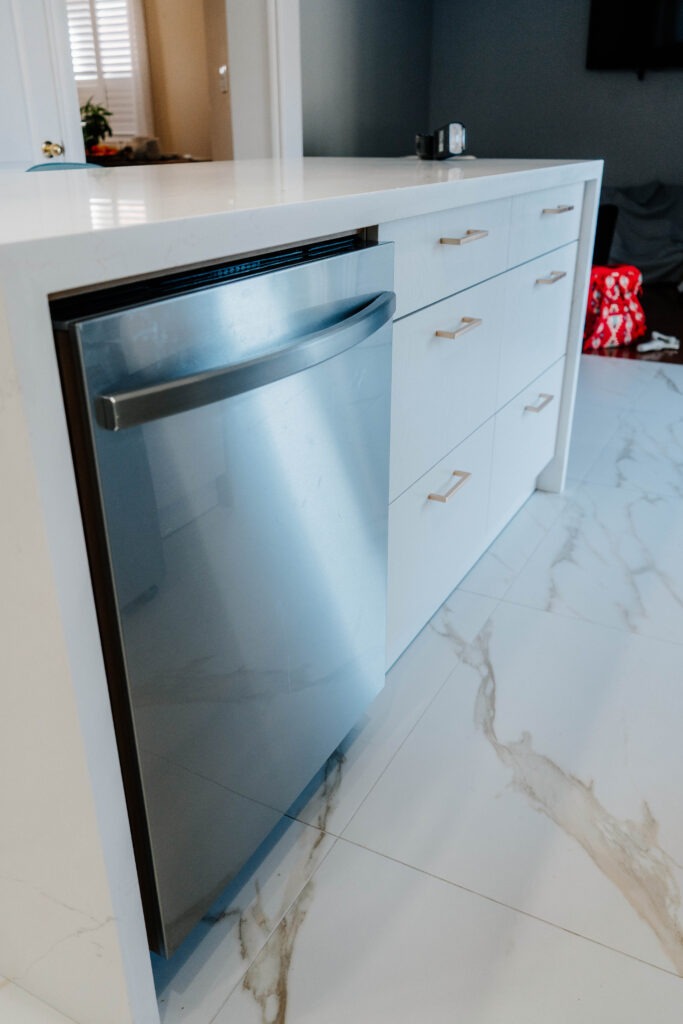 The kitchen island features an integrated stainless steel dishwasher next to Matte White drawers with gold pulls and a quartz waterfall edge