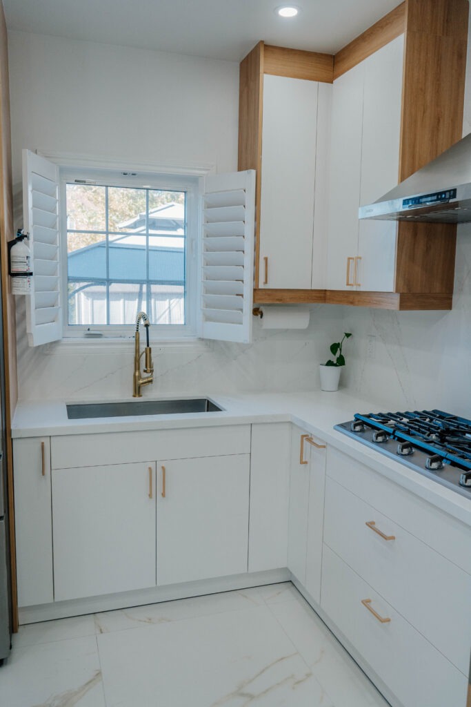 The kitchen sink area features a gold faucet and white plantation shutters, with the elegant veining of the Calacatta Gold porcelain floor visible