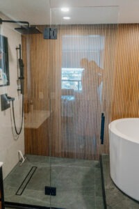 Walk in shower with frameless glass, a vertical wooden slat accent wall, and a matte black rain shower. Modern bathroom renovation, Magic Reno design