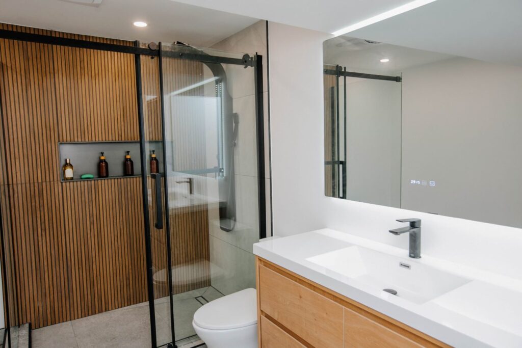 Angled view of bathroom vanity showing the matte black faucet and reflection of the wood slat shower in the LED mirror