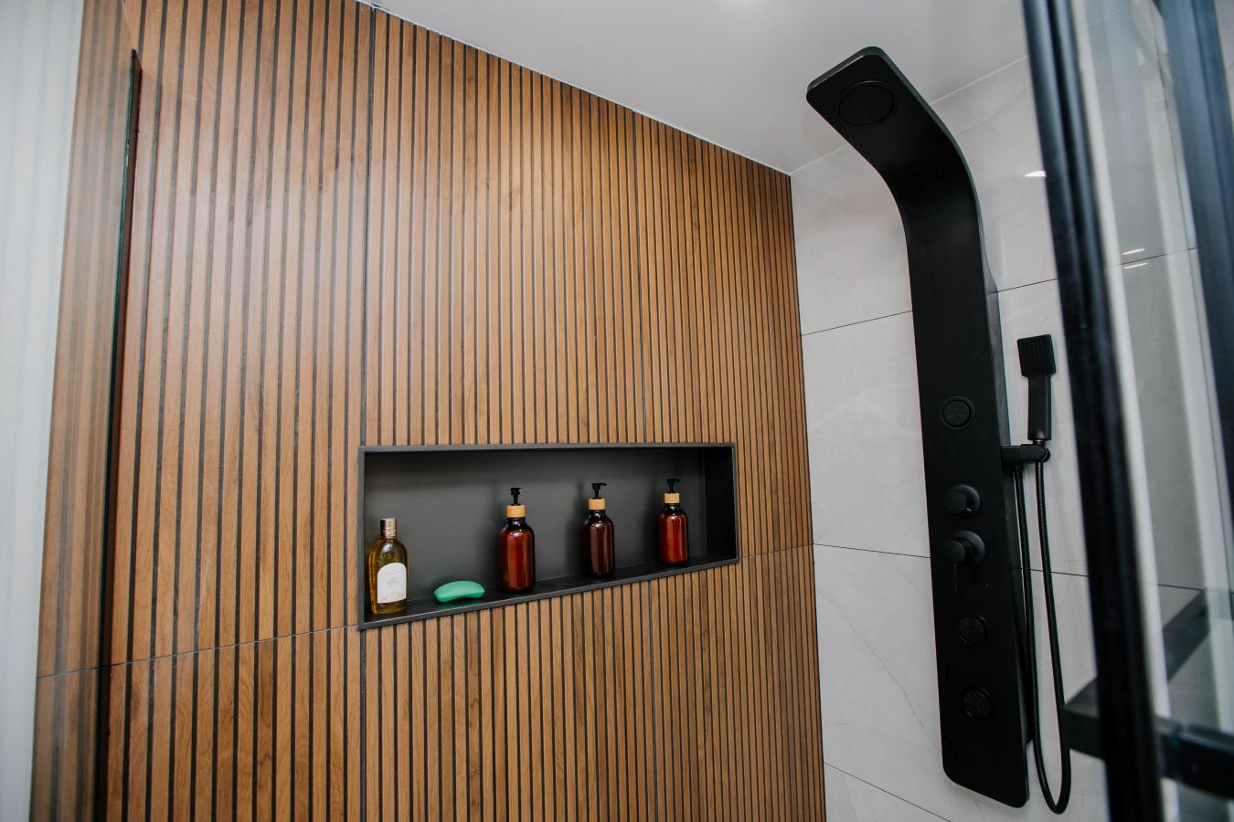 Close up of wide black recessed shower niche set into a vertical wood slat feature wall with amber soap bottles