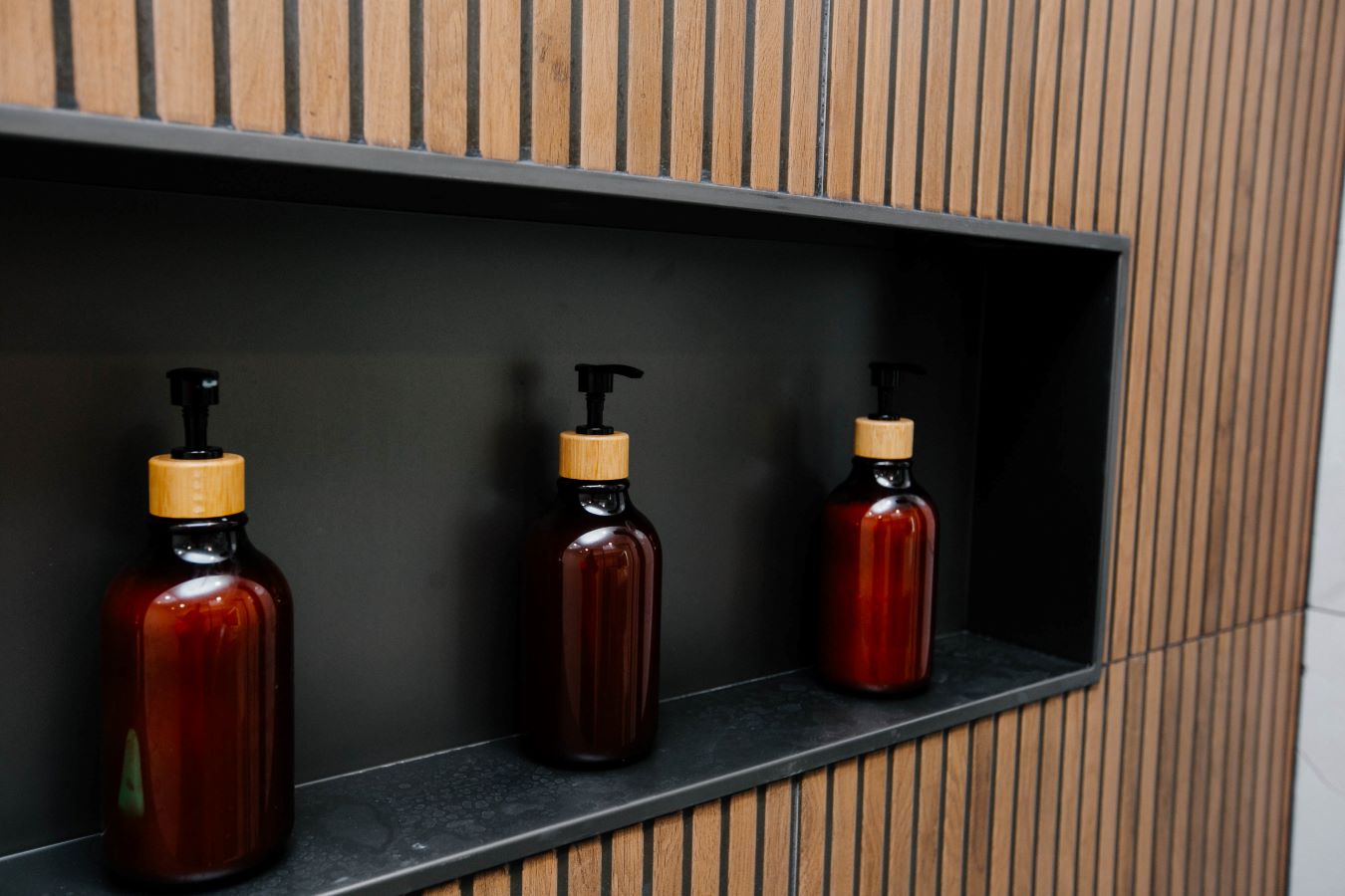 Detail shot of black shower niche shelves styled with amber glass dispenser bottles against wood slat tiling