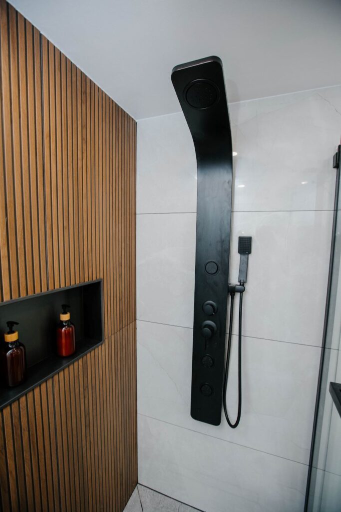 Matte black shower panel tower with rainfall head and body jets mounted on white marble look tile next to a wood slat wall