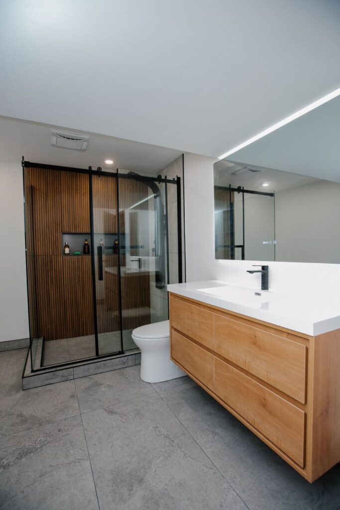 Modern bathroom featuring a vertical wood slat shower wall, black sliding glass doors, floating wood vanity with a white integrated sink, and a large LED backlit mirror