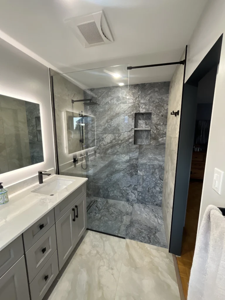 Modern grey vanity with matte black faucet and backlit mirror next to a custom glass shower with dark grey marble tiles