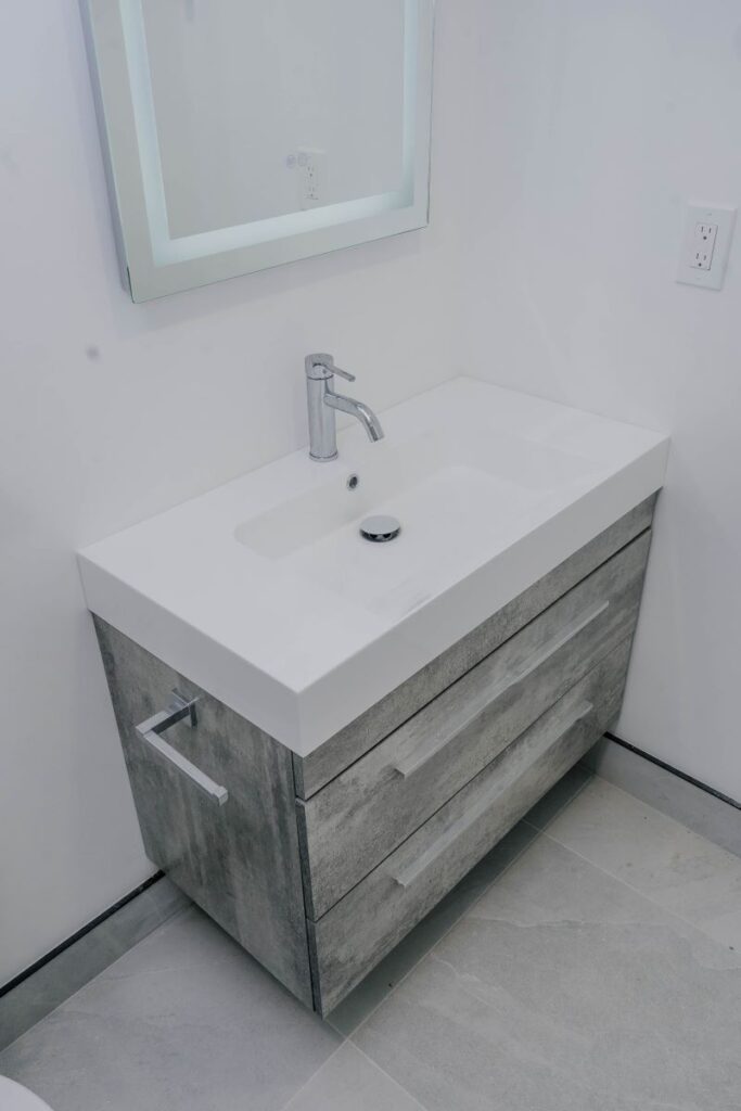 Contemporary grey wood tone vanity with a white integrated sink top, chrome faucet, and LED backlit mirror