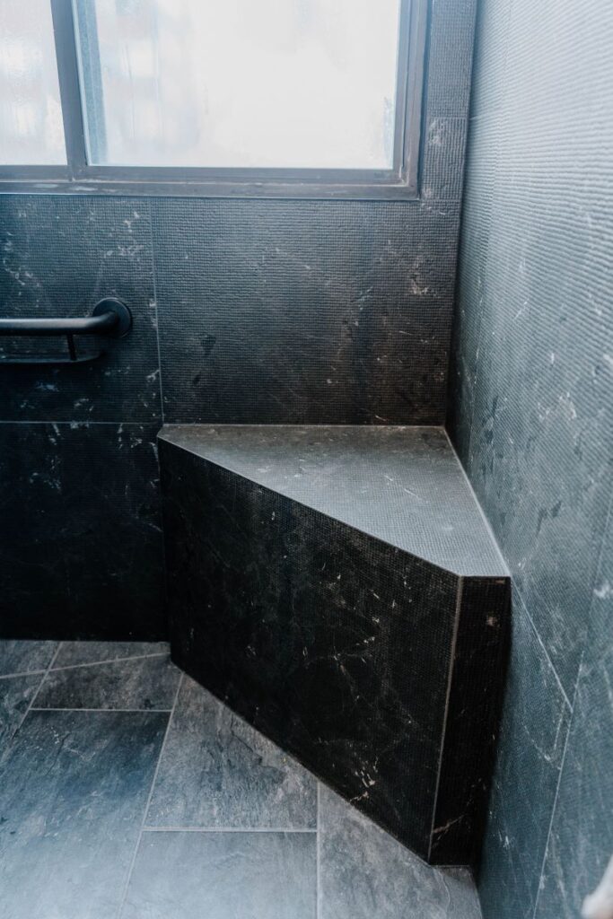 Custom built in triangular shower bench made of black stone look tile, located under a window in a high rise condo
