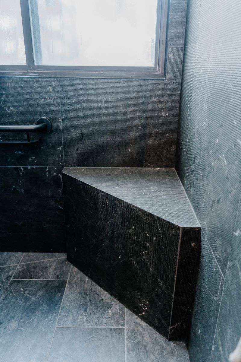 Custom built in triangular shower bench made of black stone look tile, located under a window in a high rise condo