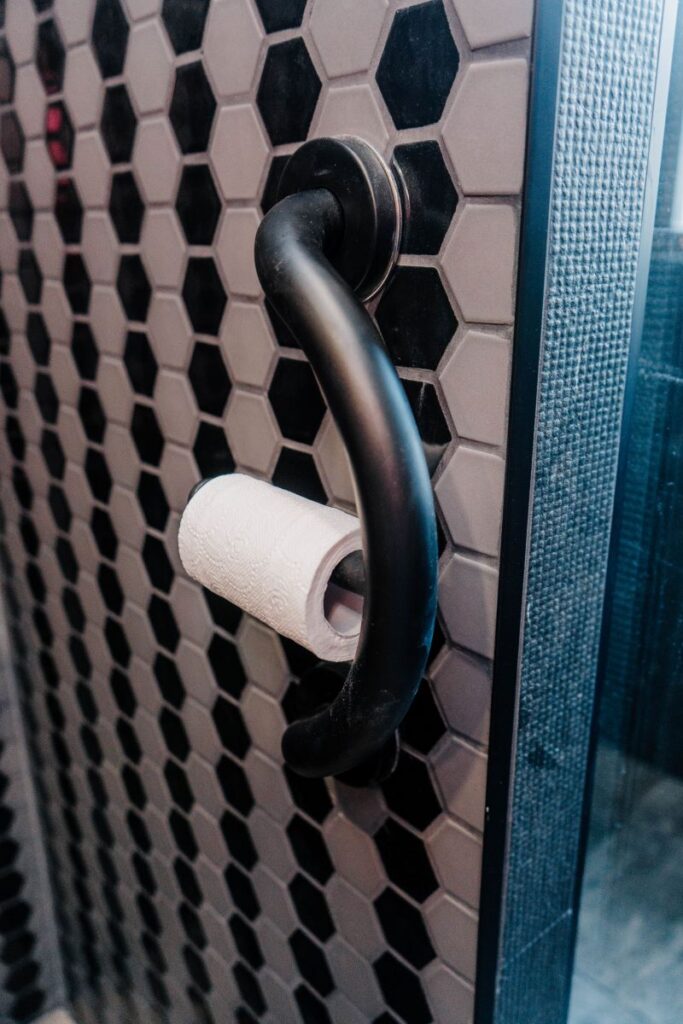Matte black toilet paper holder mounted on a custom partition wall featuring black and grey hexagon mosaic tiles