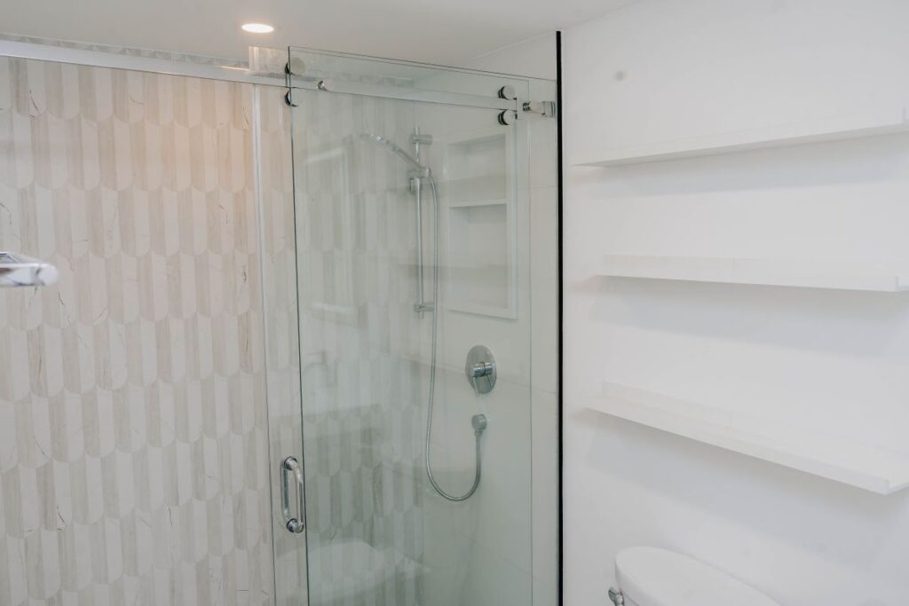 Modern condo shower renovation featuring a glass sliding door, white scallop feature tile, chrome hardware, and open shelving