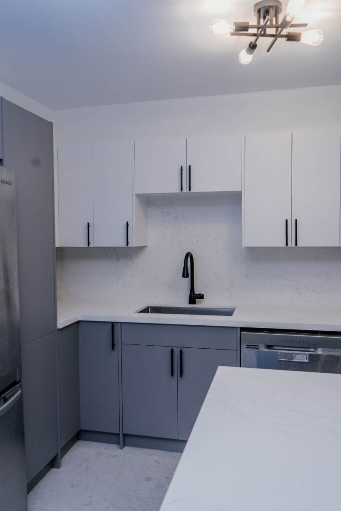 Vertical shot of a modern condo kitchen with a stainless steel fridge, grey lower cabinets, white upper cabinets, and a matte black faucet
