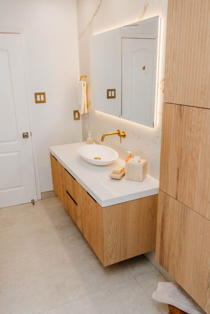 Beautifully lit modern bathroom vanity with warm oak wood tones, gold accents, and LED mirror lighting