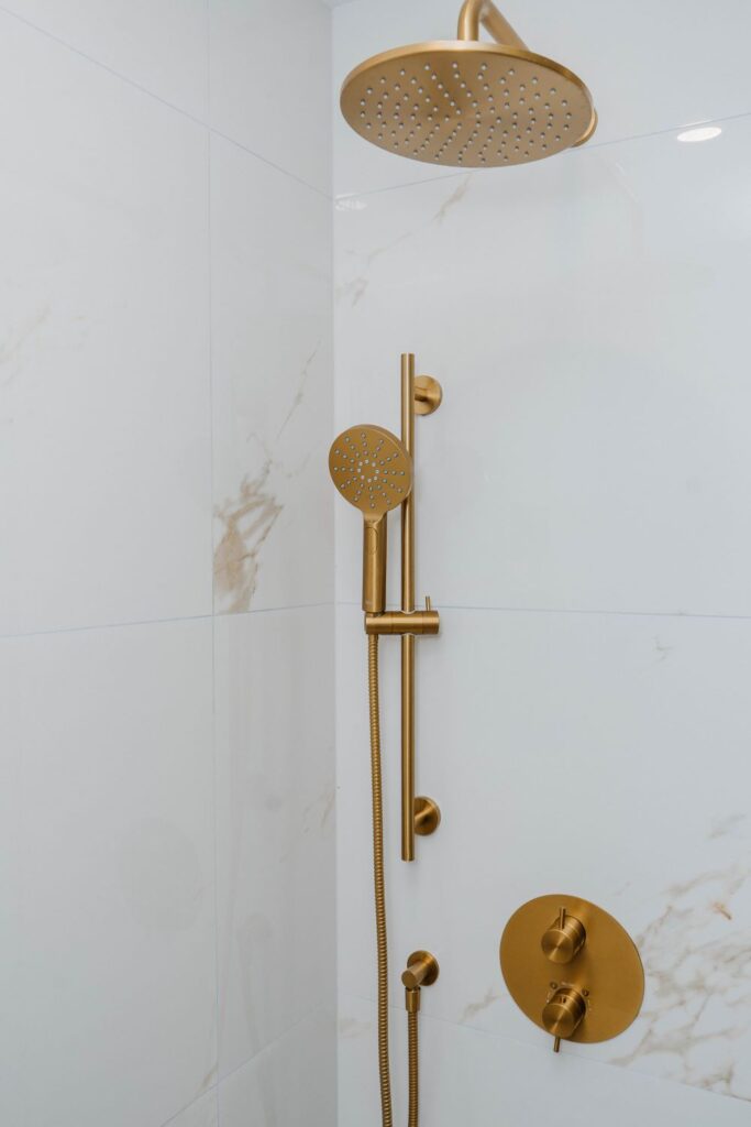 Brushed gold rain shower head and handheld wand installed against white marble look tile
