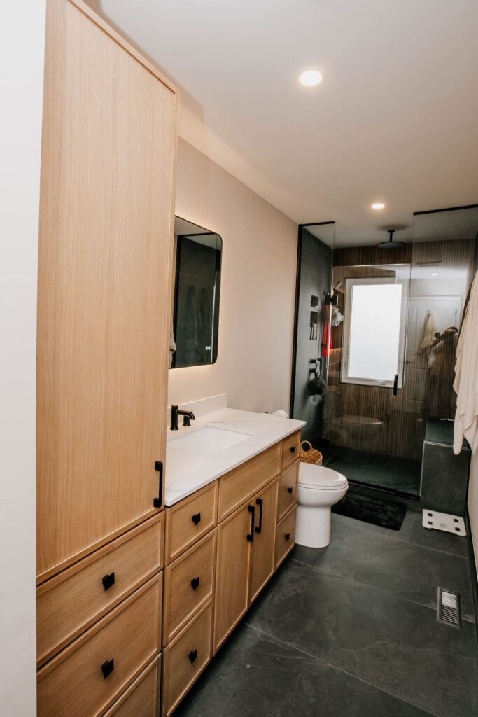 Full bathroom view featuring a light oak linen cabinet, wood vanity, and a contrasting dark walk in shower