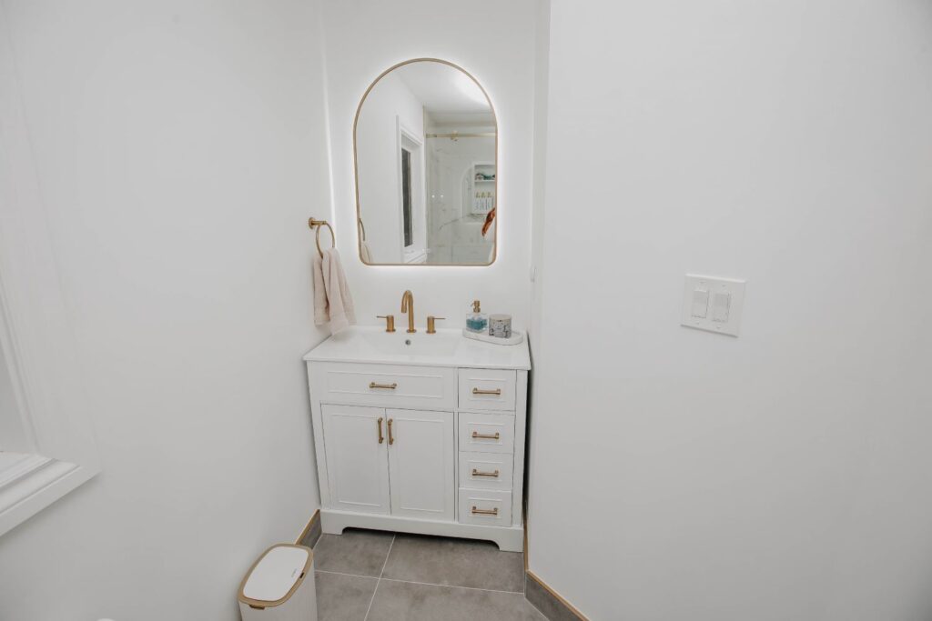 Small bathroom vanity renovation featuring white cabinetry with gold hardware, a brushed gold widespread faucet, and an arched LED mirror