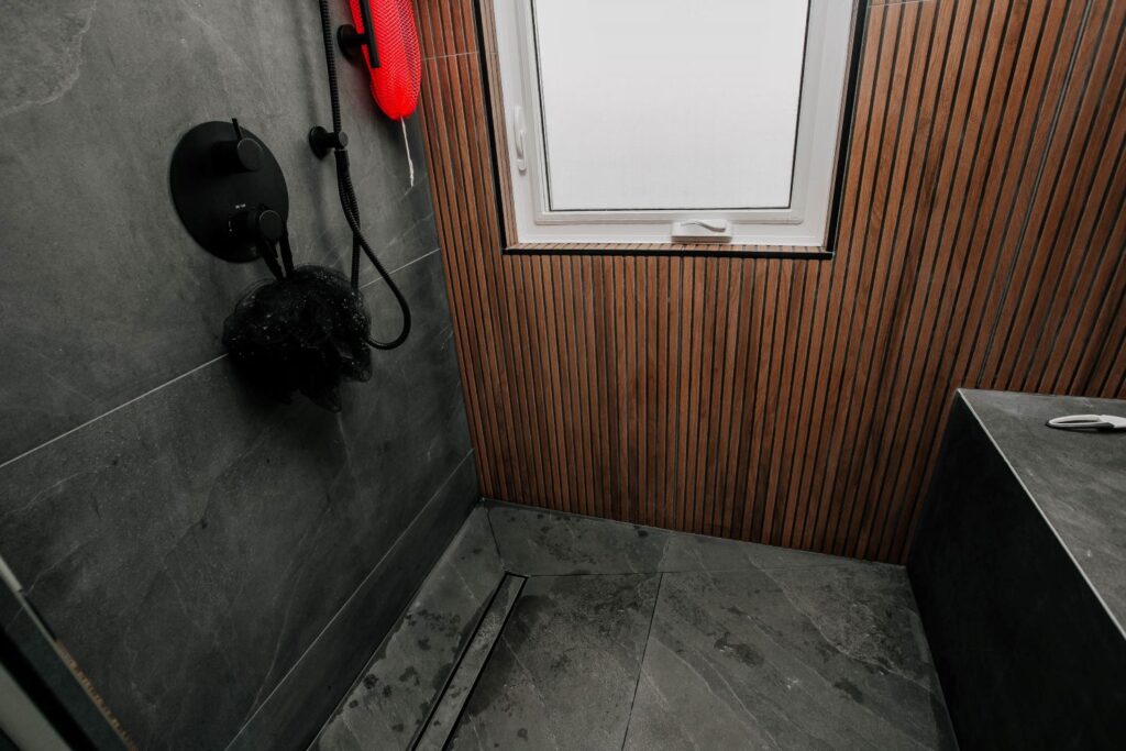 View inside a custom shower showing dark slate floor tiles, a linear drain, and a wood slat accent wall with a window