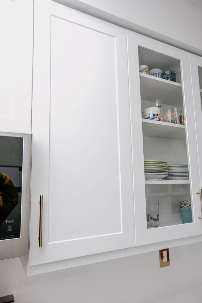 Close up of white shaker upper cabinet with glass front door showing dishware and brushed gold bar handle