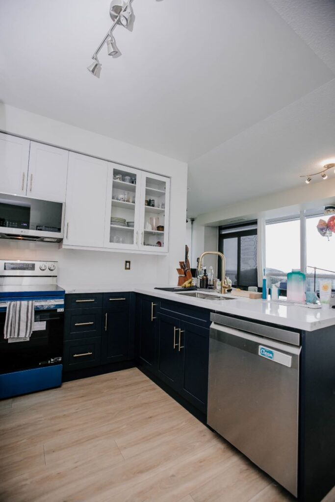 Wide view of renovated condo kitchen with navy shaker base cabinets, stainless dishwasher, quartz countertop and large window