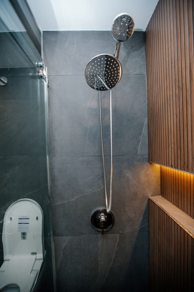 Polished chrome rain and handheld shower combo on a grey slate look wall beside a backlit oak slat feature wall